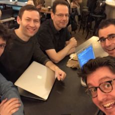 5 attendees smile at the camera while huddling around a metal table with two laptops visible.