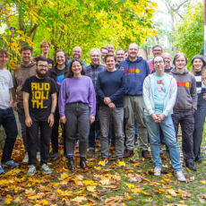 Twenty IndieWebCamp participants standing smiling roughly in 3-4 rows on an outdoors inclined driveway with yellow leaves and grass on the ground, and a canopy of trees overhead, between a mid-height retaining wall and building.