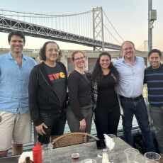 Six people standing side-by-side near an outside dining table at Red’s Java House with the Bay Bridge in the background just after sunset.