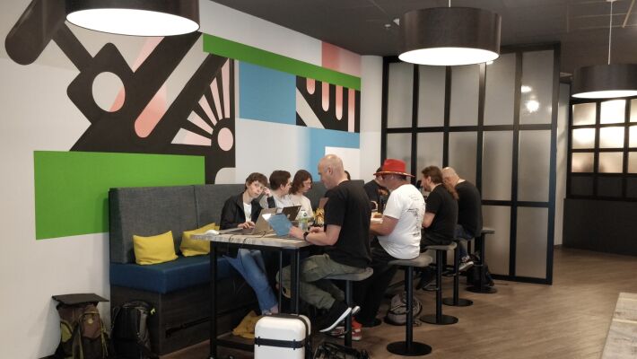 Several people using laptops sitting at a table in a spacious lobby with a large abstract artwork on the wall behind them