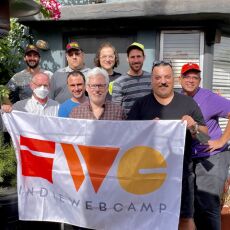 9 participants, one wearing a mouth and nose covering mask, standing outside in the sun in two staggered rows holding up a large IndieWebCamp flag, with a few plants behind them on one side.