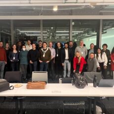 Twenty participants at the event standing side by side in roughly two rows in front of windows in an office building conference room.