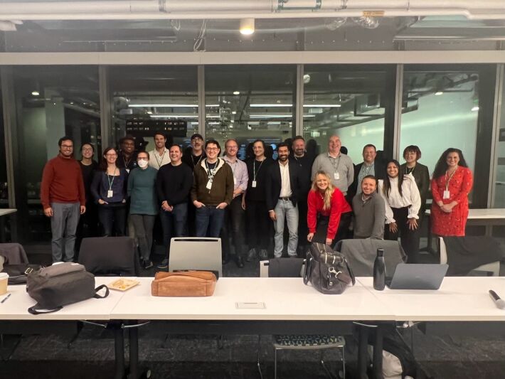 Twenty participants at the event standing side by side in roughly two rows in front of windows in an office building conference room.