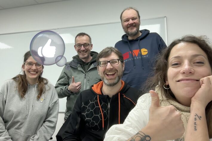 Five people smiling, sitting and standing staggered in front of a white board, with a semi-transparent graphic thumbs-up in a bubble seemingly originating from a thumbs-up gesture of one of the people.