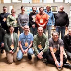 17 people gathered inside an office space smiling for a group photo.