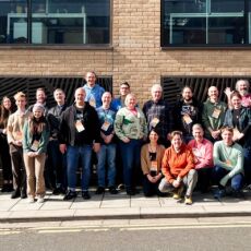 26 people gathered outside a building smiling for a group photo in the sun.