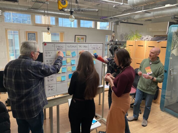 5 people gather around a whiteboard moving sticky notes as they plan a schedule.