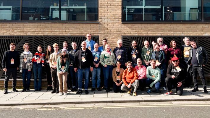 26 people gathered outside a building smiling for a group photo in the sun.