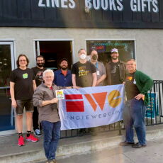 8 people standing in front of a storefront, with two of them holding a large banner printed with the logo for IWC and the word INDIEWEBCAMP.