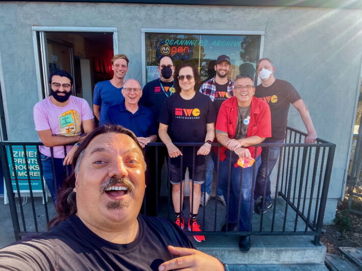 Man taking a selfie in the foreground, with 8 people standing in front of a storefront in the background.